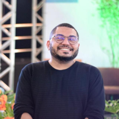 Uma pessoa de óculos e barba, vestindo um suéter preto, sorri enquanto está sentada em um ambiente interno com plantas e estruturas metálicas ao fundo, capturada em um estilo casual de Rascunho automático.