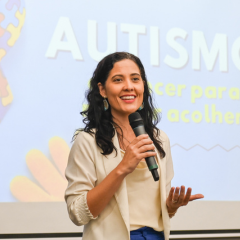 Uma mulher segurando um microfone fala em frente a uma tela que exibe a palavra "AUTISMO", texto adicional, gráficos e a frase "Rascunho automático".