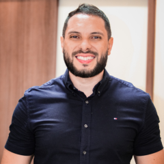 Um homem com cabelo escuro curto e barba está sorrindo, vestindo uma camisa azul marinho de botões, em pé em frente a uma porta de madeira, capturado em um momento casual de rascunho automático.