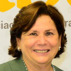 Uma mulher de meia-idade com cabelos castanhos curtos sorri para a câmera, vestindo uma blusa verde e brincos de pérola, em frente a um fundo claro com texto parcialmente amarelo e "Rascunho automático".