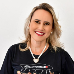Uma mulher de cabelos loiros sorri para a câmera, vestindo uma camisa preta, colar de pérolas e brincos de pérolas, em pé contra um fundo branco liso, segurando um rascunho automático.