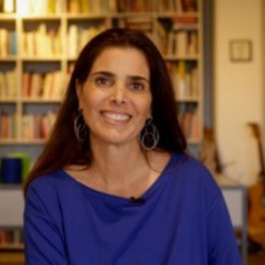 Mulher com longos cabelos castanhos, vestindo uma blusa azul, sentada em um ambiente fechado, sorrindo para a câmera, com estantes de livros e um violão ao fundo — uma cena convidativa que captura um momento de rascunho automático.