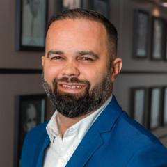 Um homem com barba e cabelo curto, vestindo um blazer azul e uma camisa branca, está em pé em frente a uma parede com fotos emolduradas, capturadas em um momento espontâneo que parece um rascunho automático.