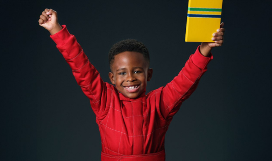 Criança sorridente, vestida de vermelho, levanta um braço e segura um livro amarelo sobre um fundo escuro, capturando a emoção da Nova Campanha.