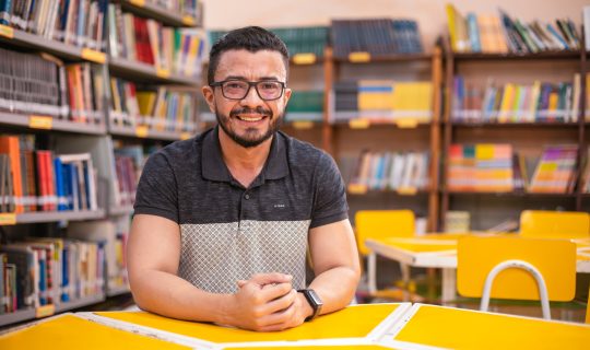 Um homem de óculos e barba senta-se em uma mesa amarela em uma biblioteca, cercado por estantes cheias de livros e informações sobre cursos.