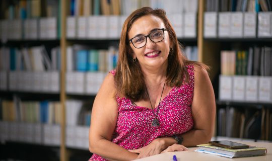 Uma mulher usando óculos e uma blusa sem mangas com estampa rosa está sentada em uma mesa com livros, um smartphone e materiais de cursos, sorrindo em frente a prateleiras cheias de arquivos e livros.