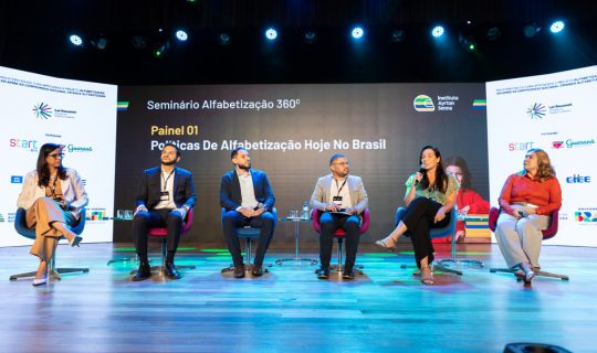 Seis painelistas sentados no palco discutindo políticas de alfabetização no Brasil no Seminário Alfabetização 360º, com a marca do evento visível ao fundo.