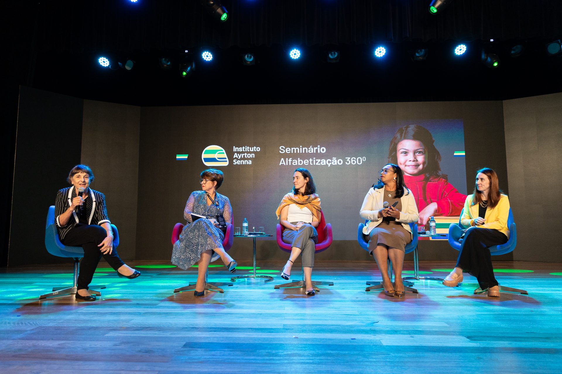 Cinco mulheres sentadas no palco em um painel de discussão no Seminário Alfabetização 360º, organizado pelo Instituto Ayrton Senna, com um telão exibindo o logotipo do evento e um retrato ao fundo.