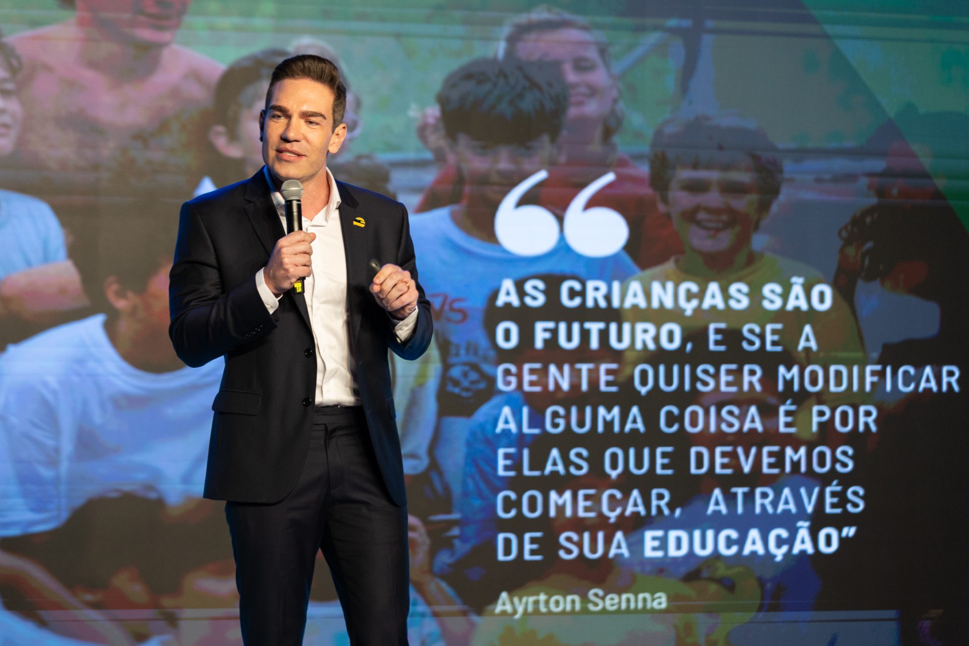 Um homem de terno fala no palco com um microfone no Seminário Alfabetização 360º. Atrás dele, um telão exibe uma frase de Ayrton Senna sobre a educação das crianças e o futuro.