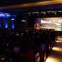 Um palestrante se apresenta no palco do Seminário Alfabetização 360º para um público sentado em um auditório pouco iluminado, com uma tela grande exibindo imagens abstratas atrás dele.