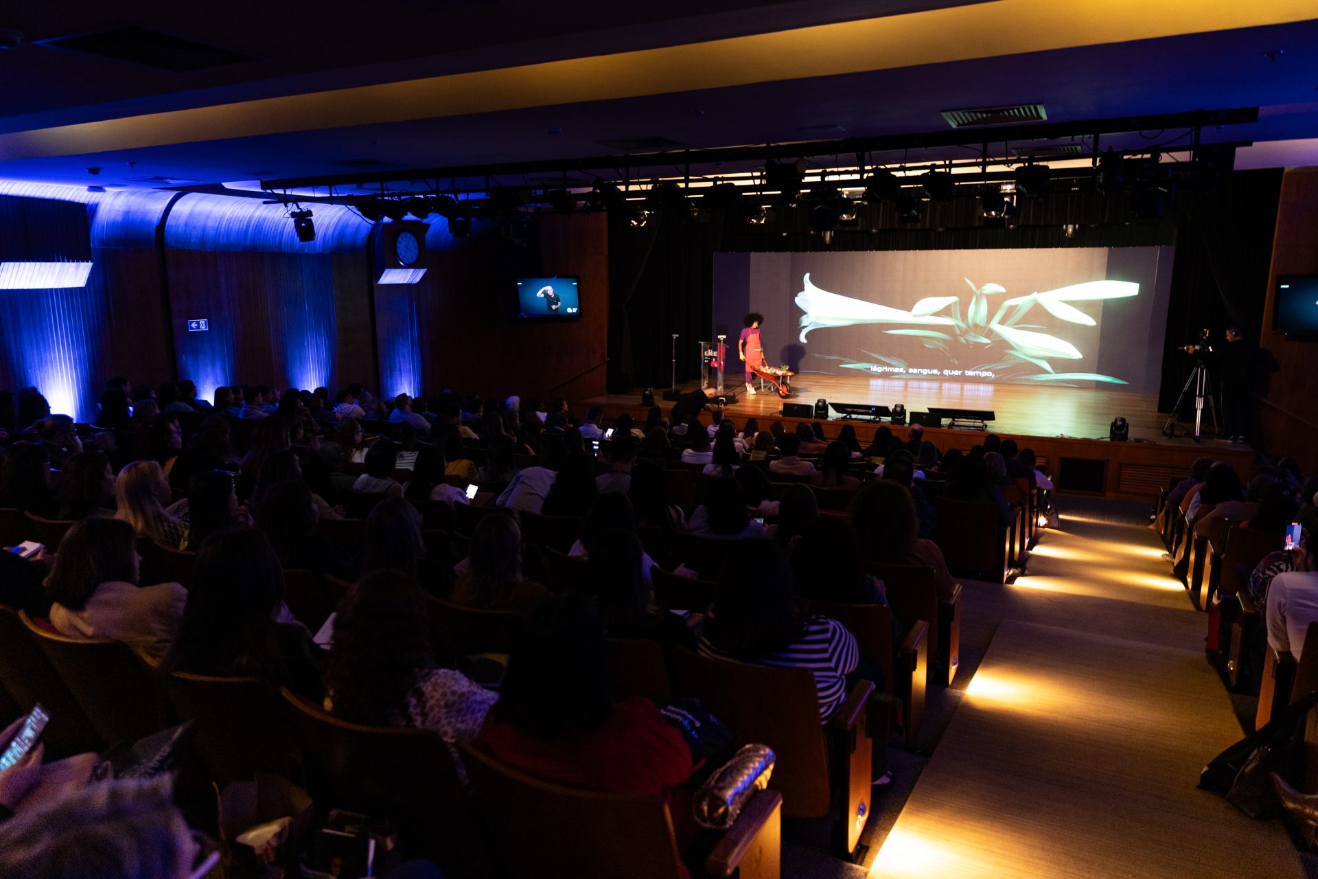 Um palestrante se apresenta no palco do Seminário Alfabetização 360º para um público sentado em um auditório pouco iluminado, com uma tela grande exibindo imagens abstratas atrás dele.