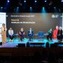 Seis palestrantes, cinco sentados e um em pé com microfone, participam do Seminário Alfabetização 360º em um palco com uma tela de apresentação e logotipos de patrocinadores ao fundo.