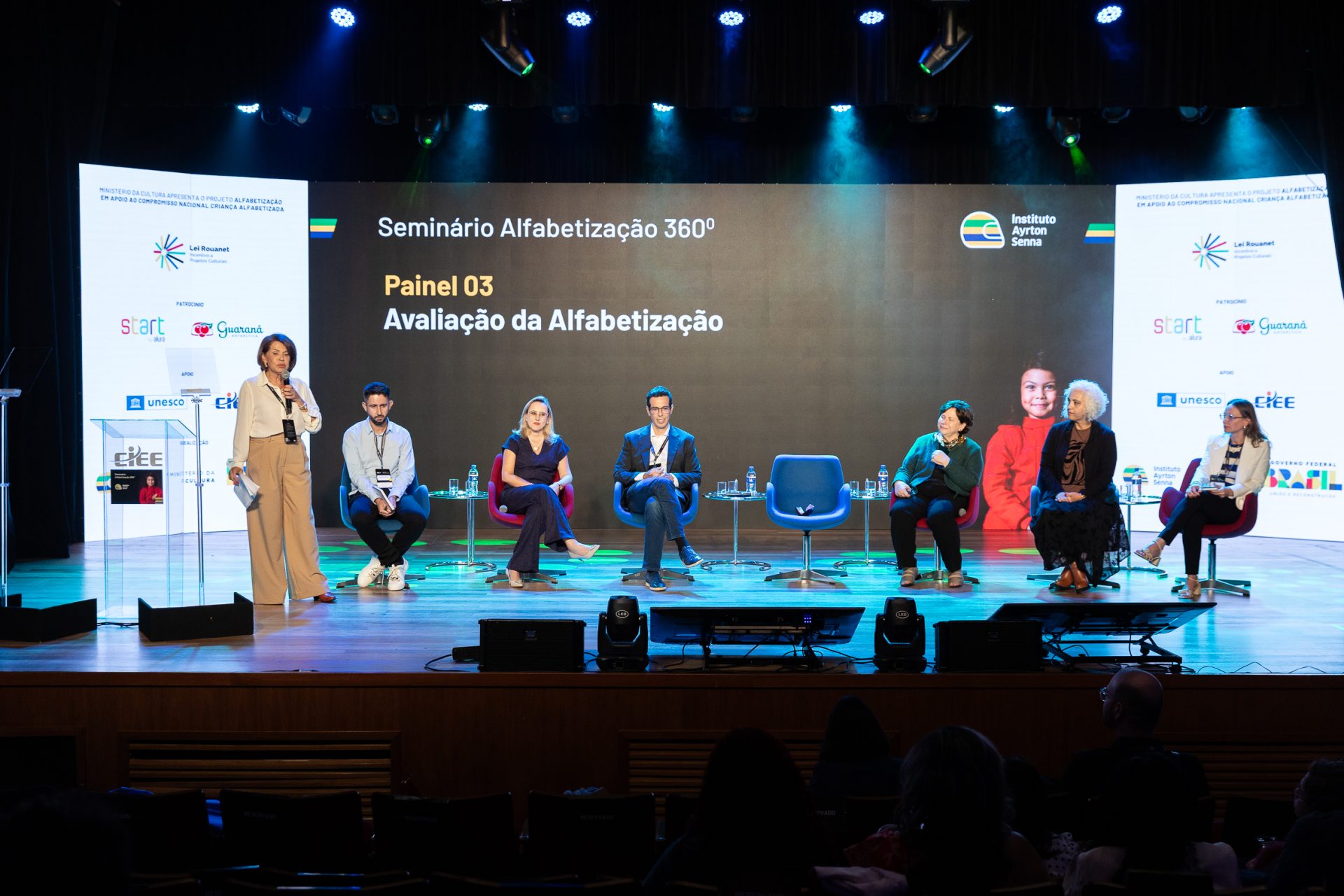 Seis palestrantes, cinco sentados e um em pé com microfone, participam do Seminário Alfabetização 360º em um palco com uma tela de apresentação e logotipos de patrocinadores ao fundo.