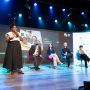Uma mulher fala no palco do painel de discussão do Seminário Alfabetização 360º, acompanhada por cinco participantes sentados com uma tela grande exibindo informações sobre o evento atrás deles.