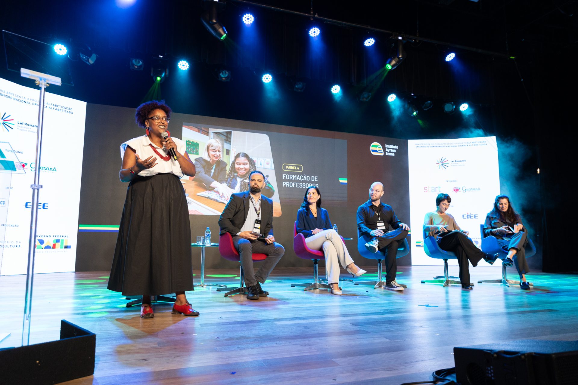 Uma mulher fala no palco do painel de discussão do Seminário Alfabetização 360º, acompanhada por cinco participantes sentados com uma tela grande exibindo informações sobre o evento atrás deles.