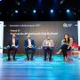 Seis painelistas sentam-se em um palco sob luzes brilhantes no Seminário Alfabetização 360º, participando de uma discussão animada sobre políticas de alfabetização no Brasil. Um grande display atrás deles destaca os principais detalhes do evento Seminário Alfabetização 360º.