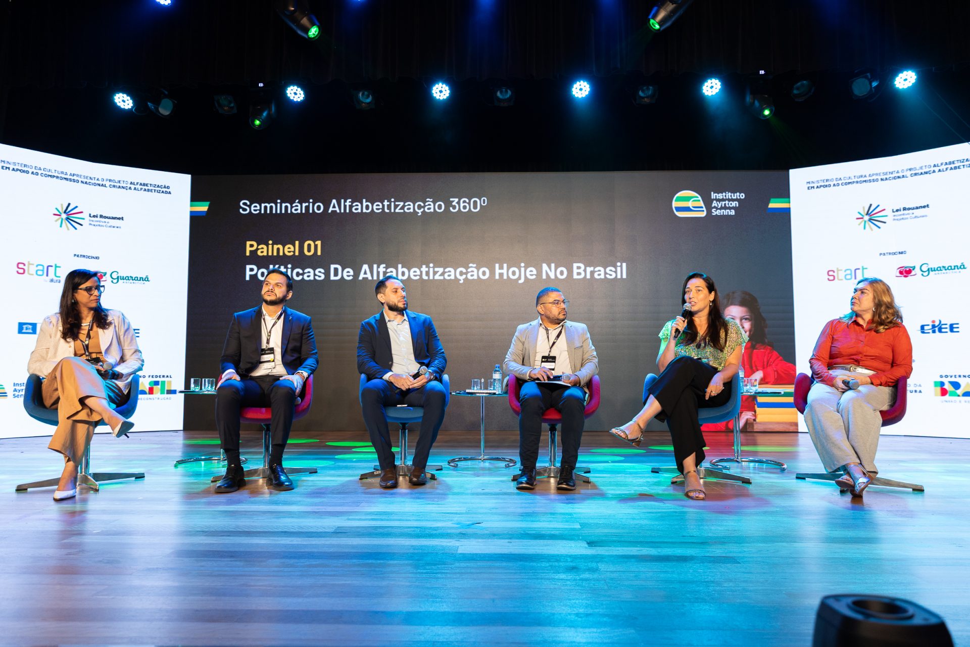 Seis painelistas sentam-se em um palco sob luzes brilhantes no Seminário Alfabetização 360º, participando de uma discussão animada sobre políticas de alfabetização no Brasil. Um grande display atrás deles destaca os principais detalhes do evento Seminário Alfabetização 360º.