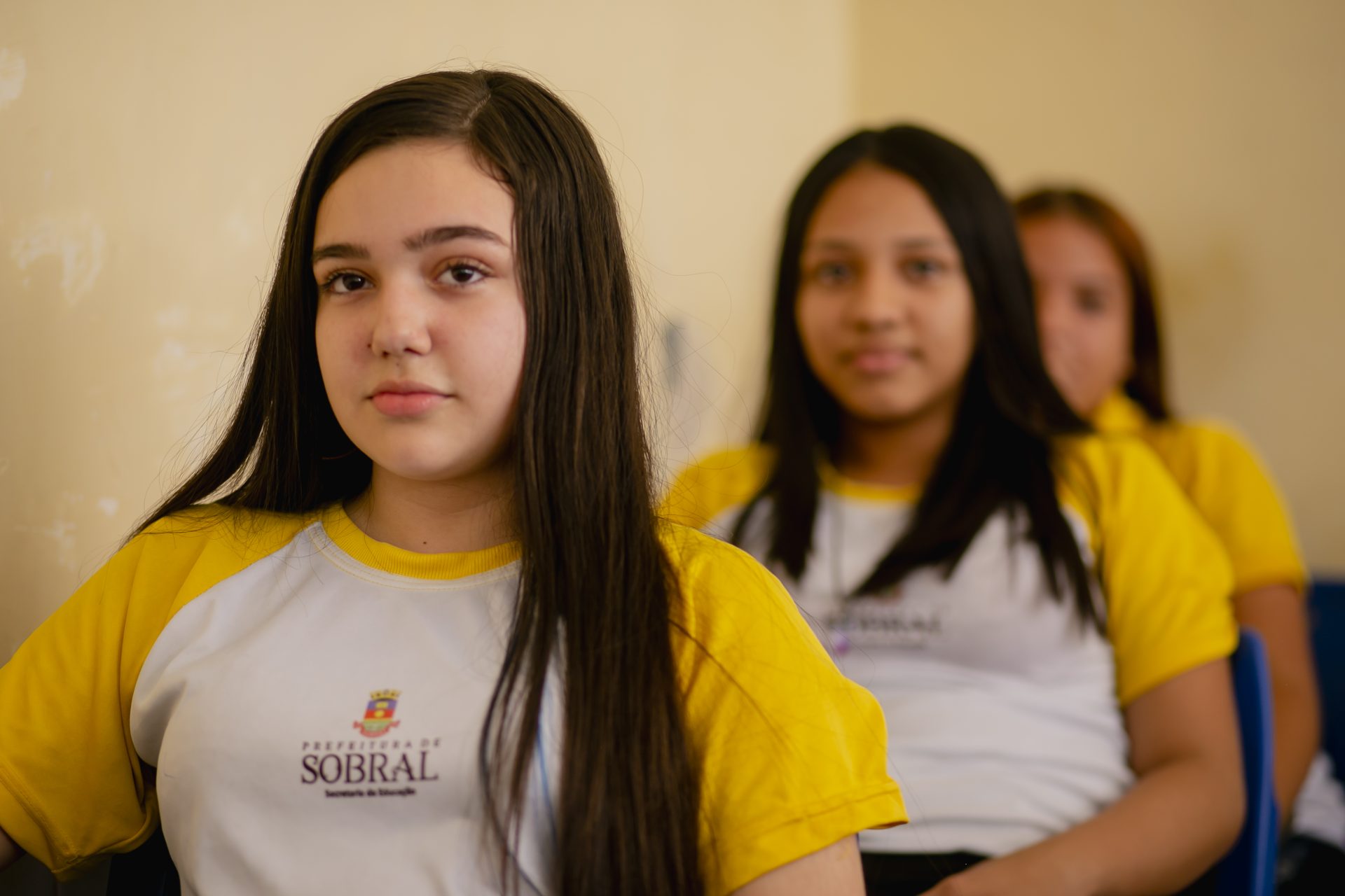 Quatro alunas usando uniformes escolares amarelos e brancos sentadas em uma sala de aula, voltadas para a frente; o foco está na aluna mais próxima da câmera durante um evento especial do McDia Feliz.