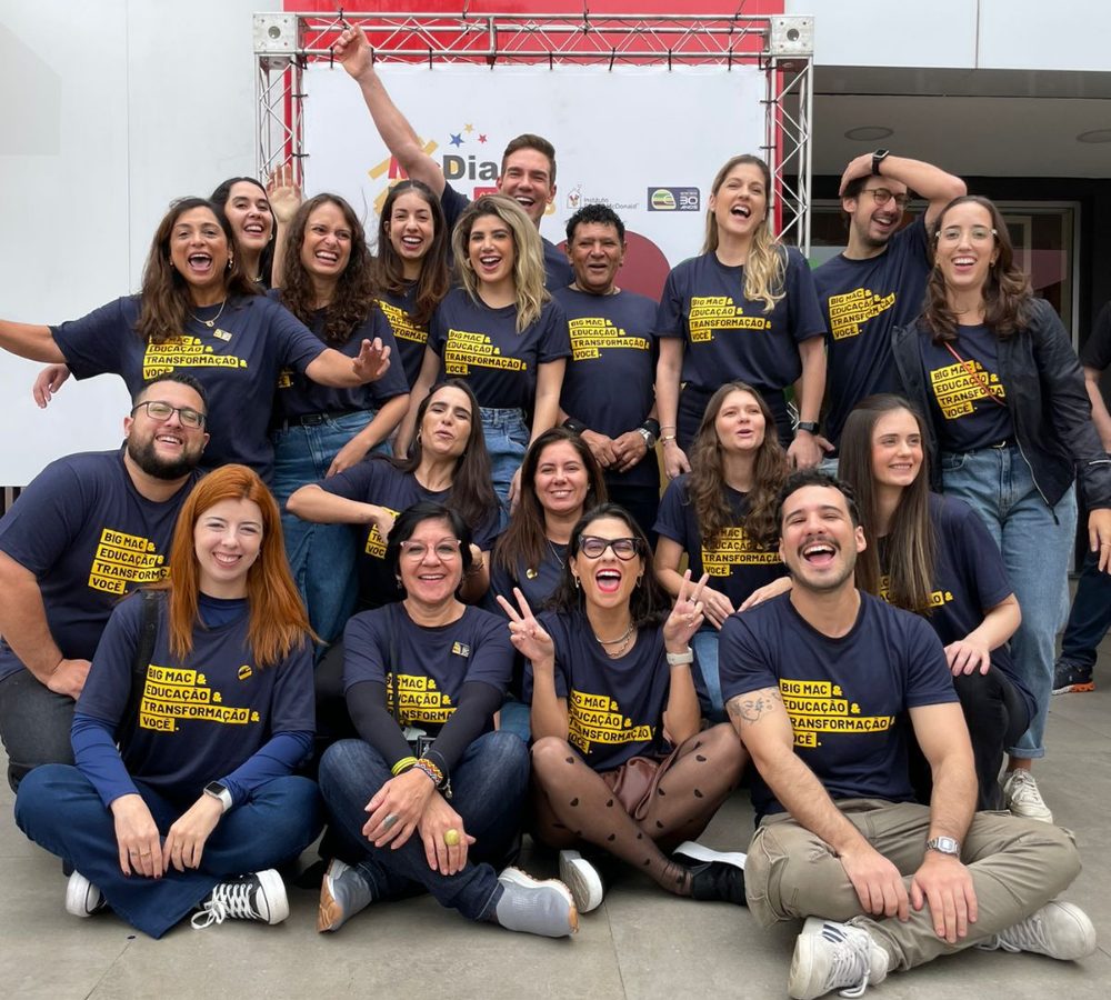 Um grupo de pessoas vestindo camisetas escuras iguais posa e sorri para uma foto em grupo em frente a um cartaz e uma moldura de metal vermelha durante o McDia Feliz 2024.