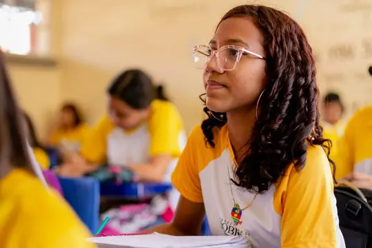 Um aluno com óculos senta-se em uma mesa em uma sala de aula, ouvindo atentamente e fazendo anotações durante o evento McDia Feliz. Outros alunos com uniformes amarelos e brancos podem ser vistos ao fundo.