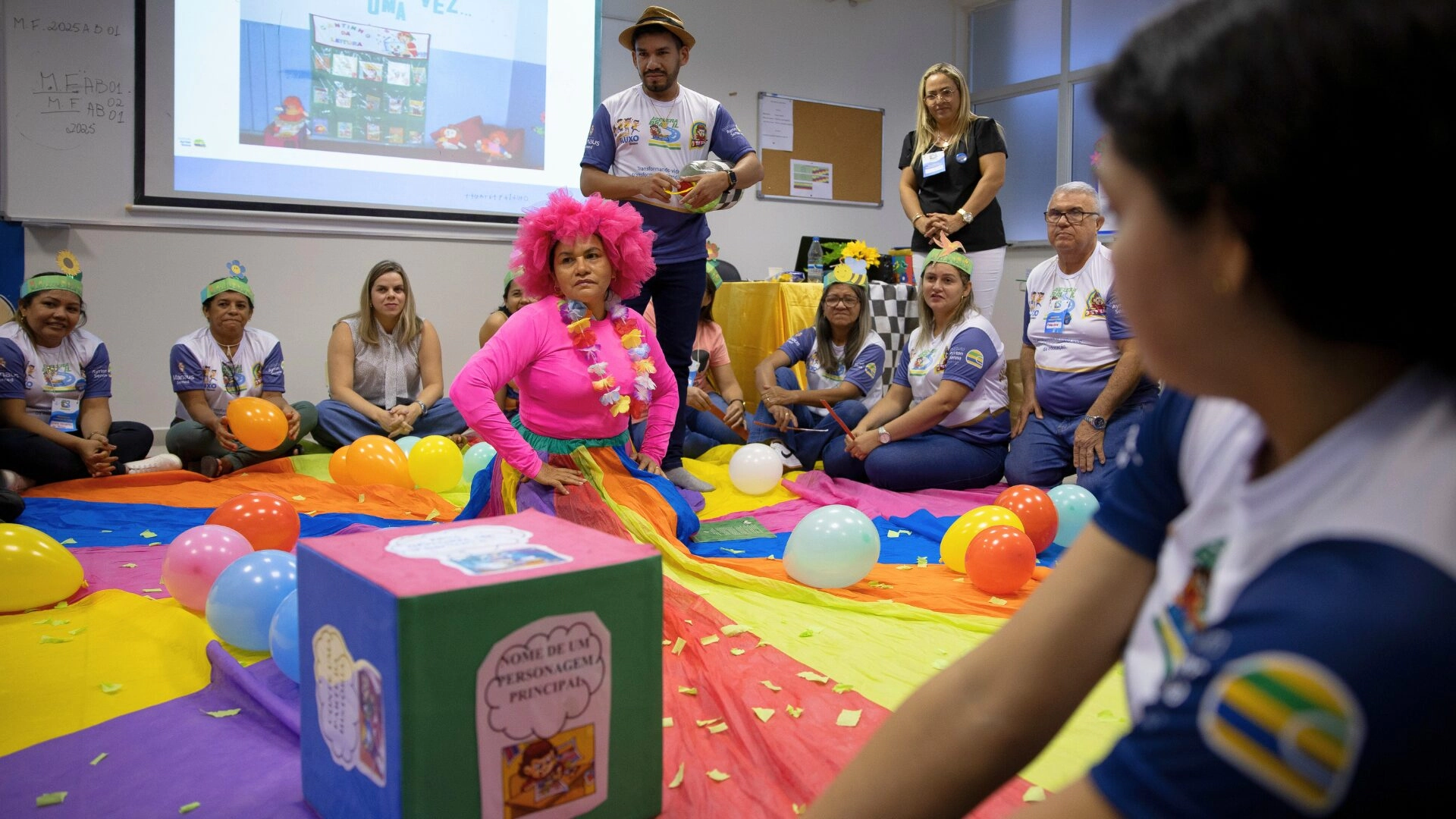Um grupo de adultos senta-se em um tapete colorido com balões e confetes, assistindo à apresentação de uma pessoa com peruca e fantasia rosa em uma sala de aula decorada para uma atividade da F1 in Schools.