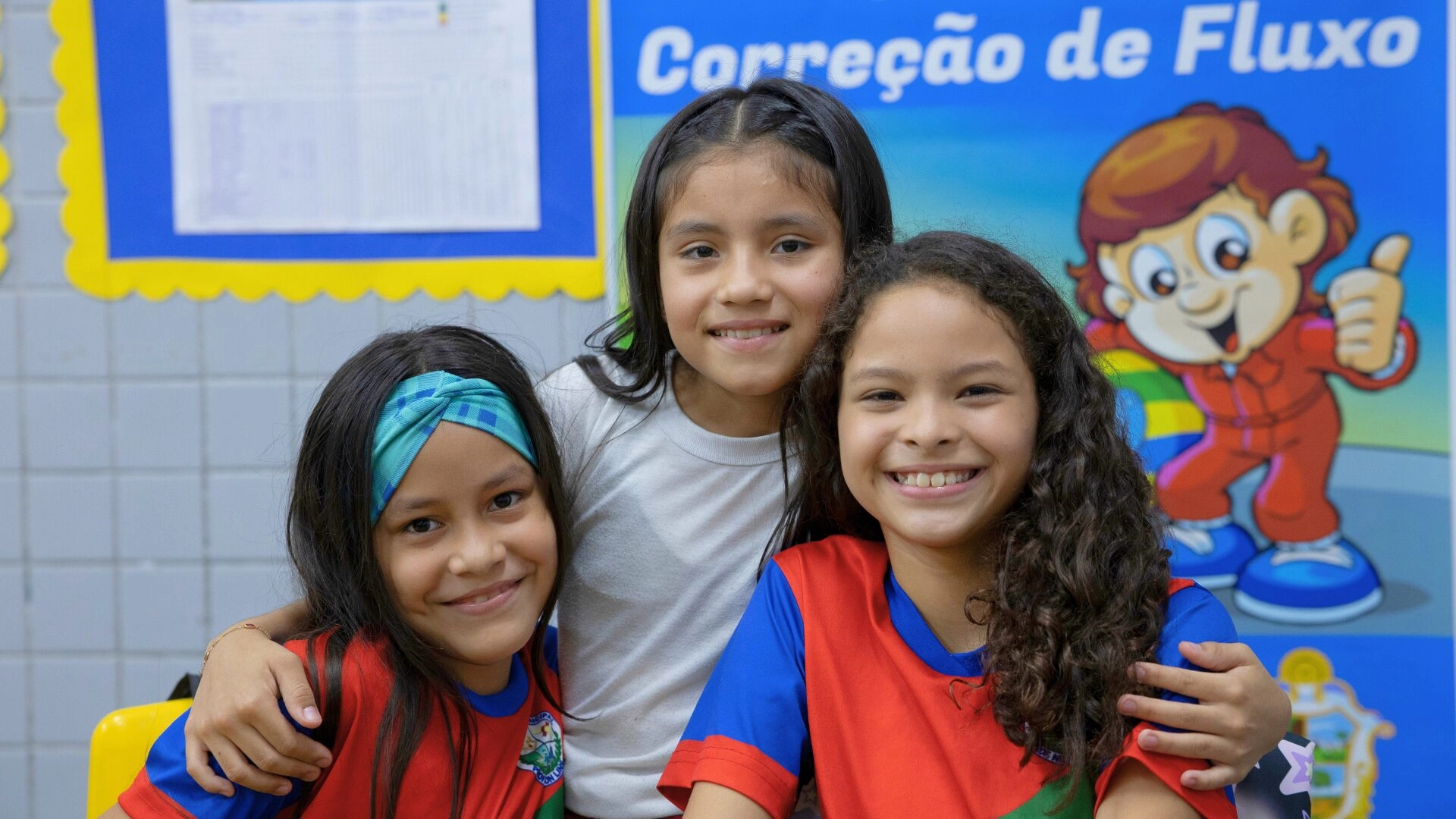 Três garotas sorridentes posam juntas em uma sala de aula, duas vestindo camisas vermelhas e azuis e uma de branco. Um pôster educacional colorido sobre F1 nas escolas pode ser visto ao fundo.