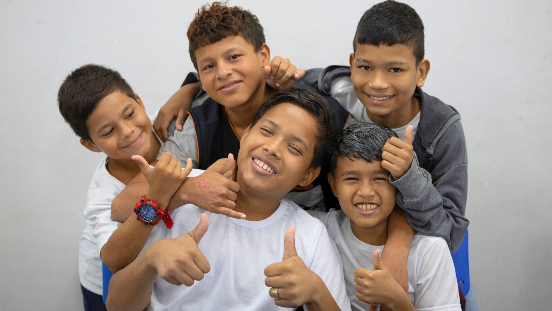 Cinco meninos posam juntos em frente a uma parede lisa, sorrindo e fazendo gestos de polegar para cima, com alguns se abraçando, representando orgulhosamente sua equipe F1 in Schools.