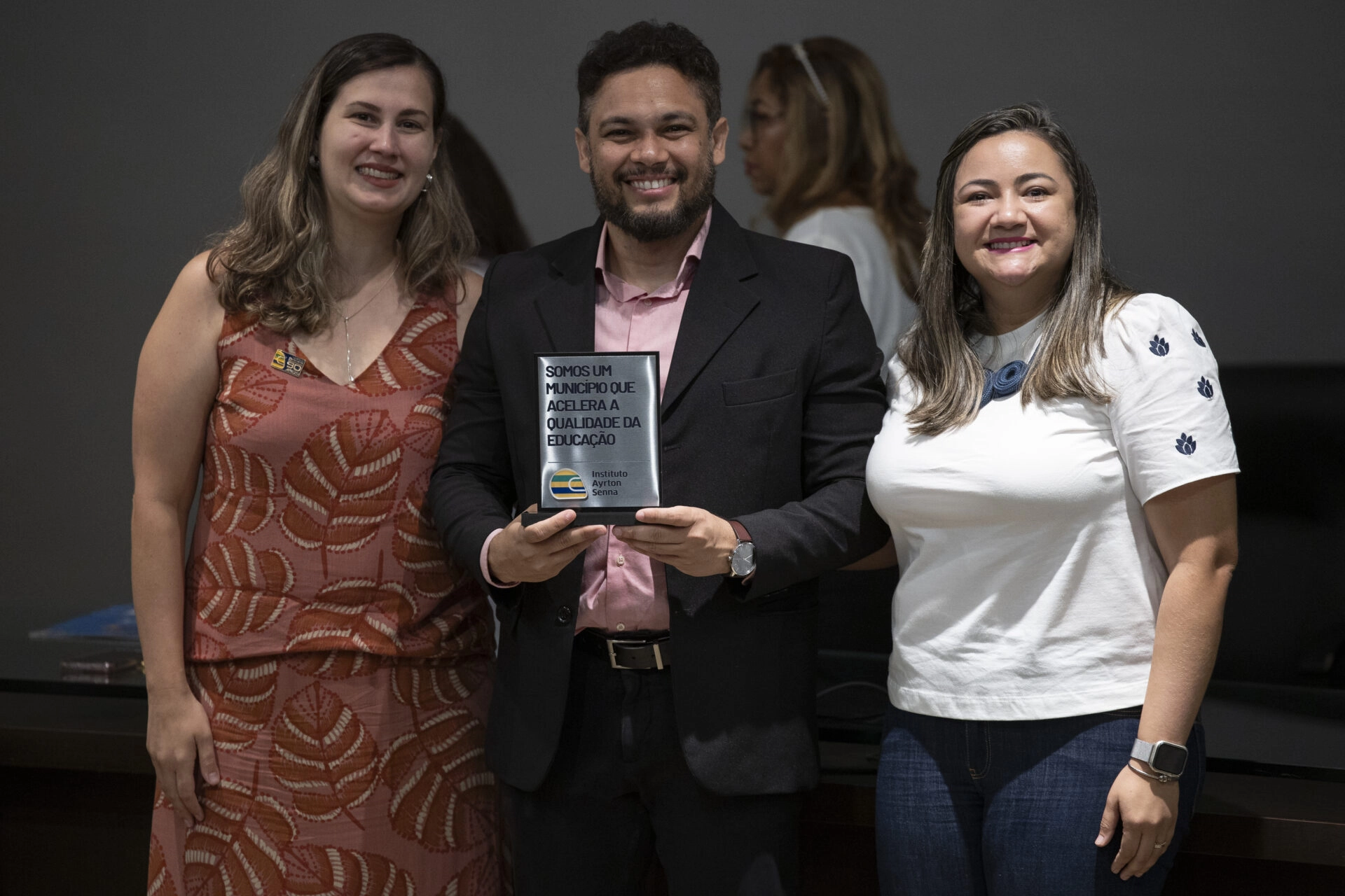 Três pessoas em pé dentro de casa, sorrindo para a câmera; a pessoa no centro segura uma placa retangular com texto em português, comemorando seu sucesso na competição F1 in Schools.