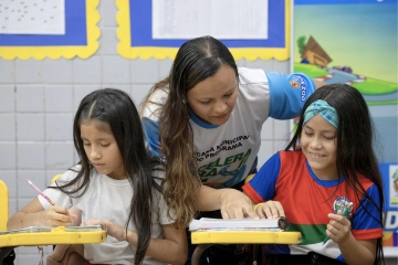Um professor auxilia duas meninas com seus trabalhos escolares em suas carteiras em uma sala de aula, apresentando a elas os fundamentos da F1 nas escolas.