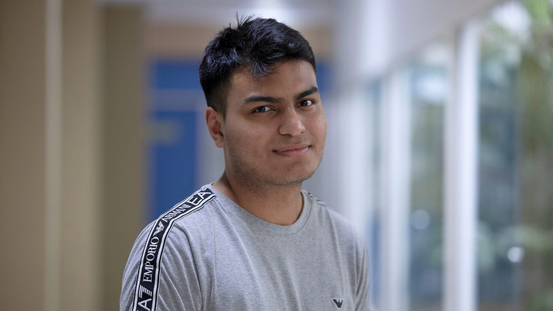 Um jovem de cabelos curtos e escuros, vestindo uma camiseta cinza da Emporio Armani, está em pé dentro de casa, olhando para a câmera com um leve sorriso, representando orgulhosamente a F1 in Schools.