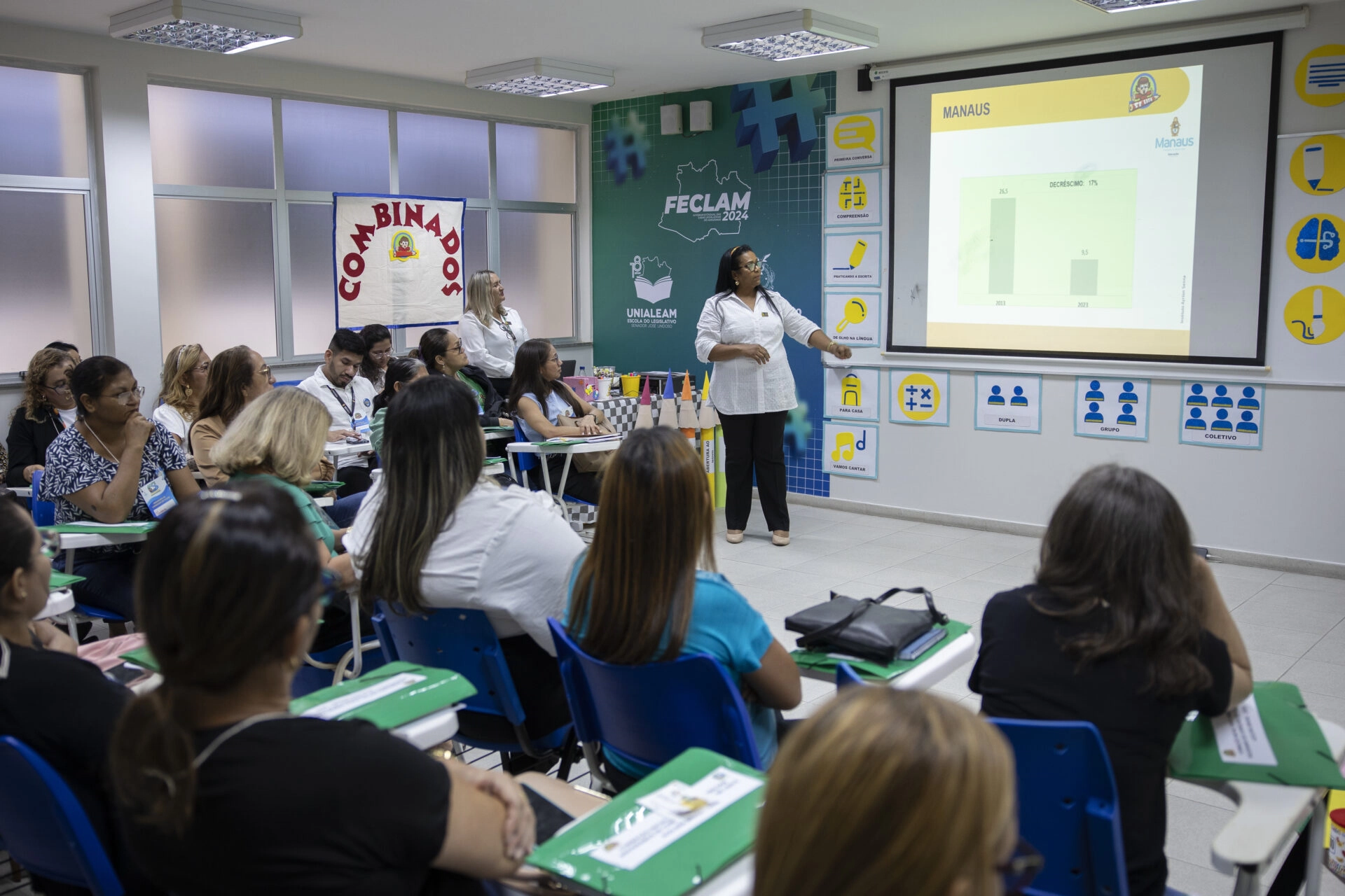 Uma mulher apresenta um gráfico de barras em uma tela de projetor para uma sala de aula com adultos sentados, que estão fazendo anotações e ouvindo atentamente durante um workshop da F1 in Schools.