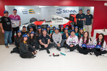 Um grupo de pessoas posa em frente a um carro antigo de Fórmula 1 no Instituto Ayrton Senna, algumas usando bonés e uniformes combinando, comemorando sua participação no programa F1 in Schools.