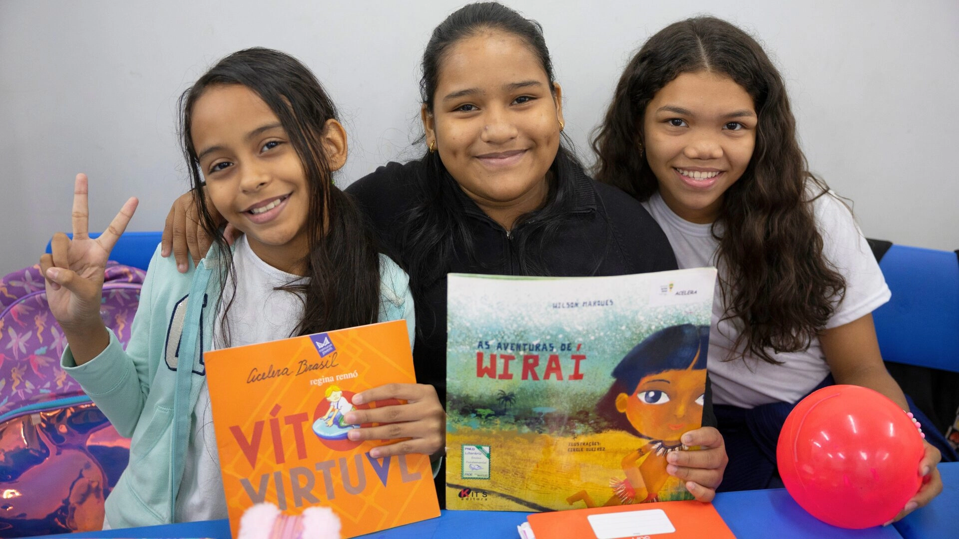 Três meninas sentadas em uma escrivaninha, sorrindo, segurando livros infantis e um balão vermelho, com material escolar e um kit do projeto F1 in Schools sobre a mesa à sua frente.