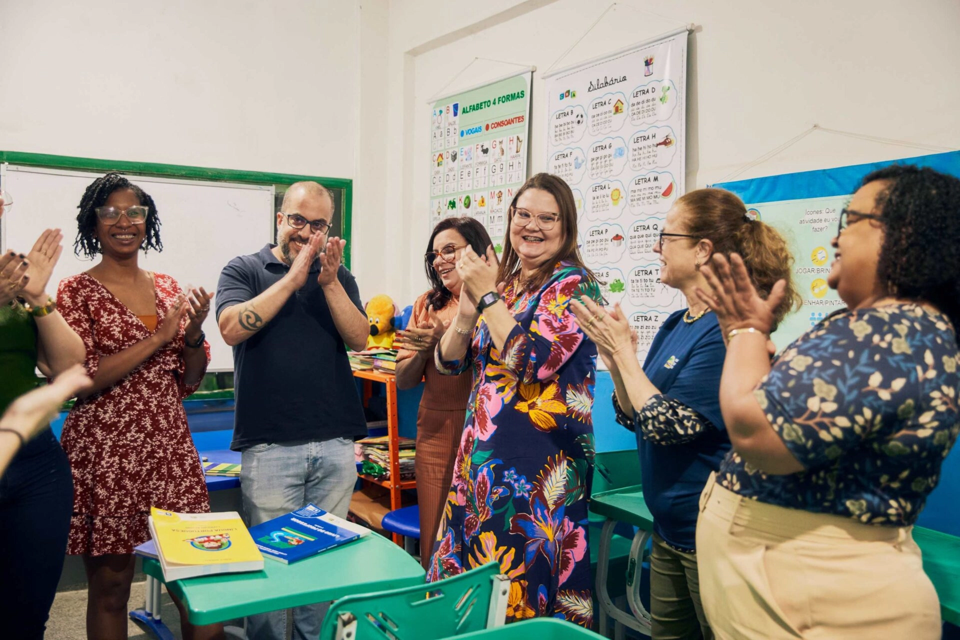 Um grupo de adultos em uma sala de aula, sorrindo e batendo palmas, com pôsteres educacionais.