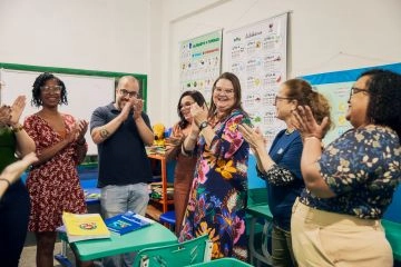 Um grupo de adultos em uma sala de aula, sorrindo e batendo palmas, com pôsteres educacionais.