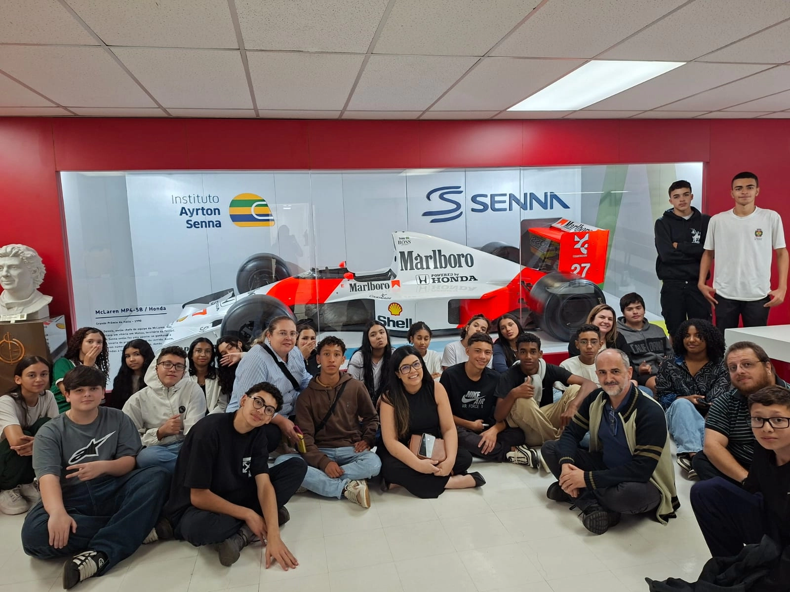 Um grupo de pessoas se senta e fica em frente a uma exibição do carro de Fórmula 1 Ayrton Senna no Instituto Ayrton Senna, com paredes vermelhas e brancas ao fundo.