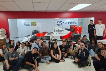 Um grupo de pessoas posa em frente a uma exposição de carros de corrida de Fórmula 1 com memorabilia de Ayrton Senna.
