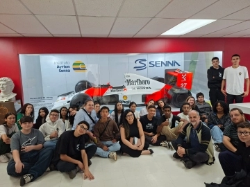 Um grupo de pessoas posa em frente a uma exposição de carros de corrida de Fórmula 1 com memorabilia de Ayrton Senna.
