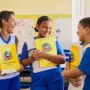 Três crianças com uniformes azuis e brancos estão em uma sala de aula, sorrindo e segurando livros didáticos amarelos de língua portuguesa do Plano Anual Instituto Ayrton Senna.