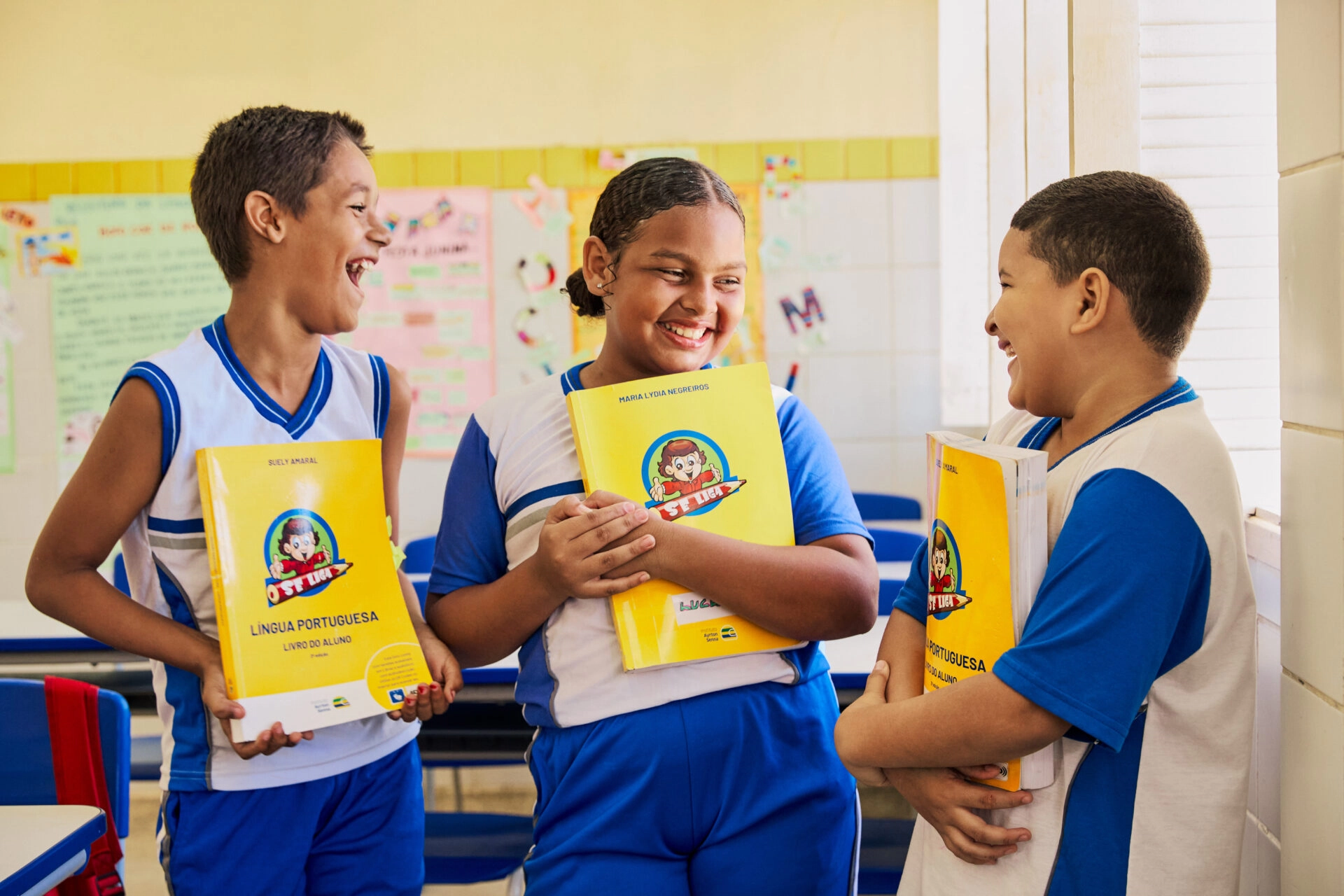 Três crianças com uniformes azuis e brancos estão em uma sala de aula, sorrindo e segurando livros didáticos amarelos de língua portuguesa do Plano Anual Instituto Ayrton Senna.