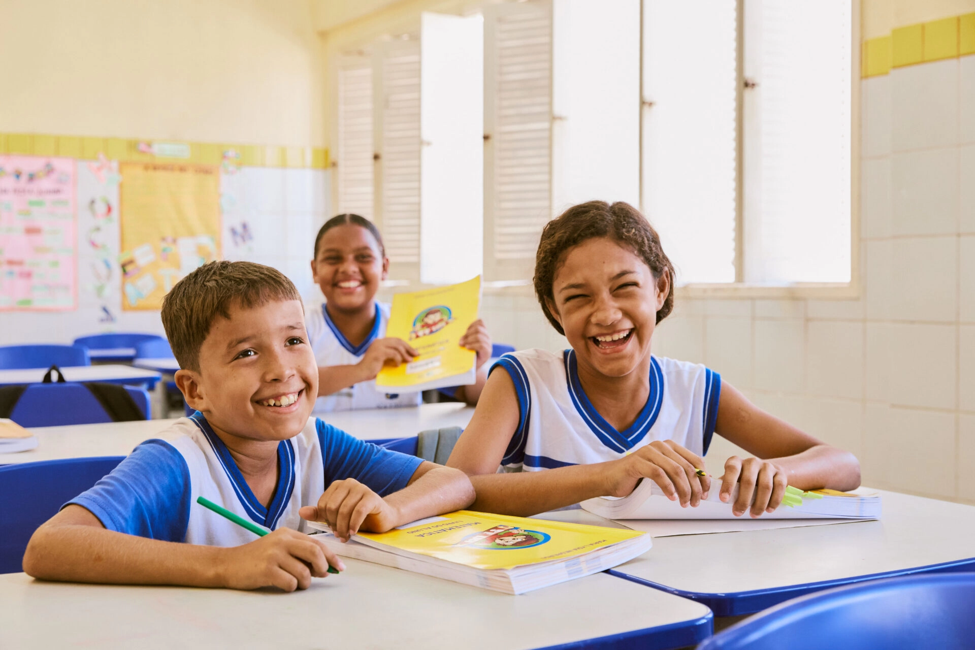 Três alunos em uma sala de aula sentados em carteiras, sorrindo e segurando livros de exercícios, usando uniformes brancos e azuis, como parte da iniciativa do Plano Anual Instituto Ayrton Senna.