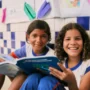 Duas crianças com uniformes escolares sentam-se no chão, sorrindo e lendo livros juntas. Atrás delas, desenhos e papéis decoram uma parede de azulejos azuis e brancos - uma cena inspiradora que reflete o impacto do Plano Anual Instituto Ayrton Senna.