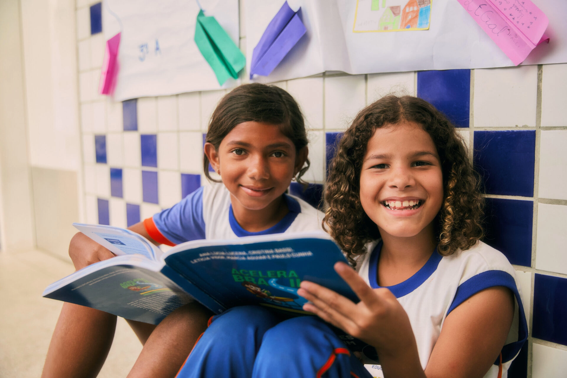 Duas crianças com uniformes escolares sentam-se no chão, sorrindo e lendo livros juntas. Atrás delas, desenhos e papéis decoram uma parede de azulejos azuis e brancos - uma cena inspiradora que reflete o impacto do Plano Anual Instituto Ayrton Senna.
