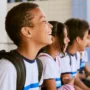 Quatro alunos usando uniformes e mochilas iguais sentam-se em uma fileira, sorrindo e olhando para a esquerda em um ambiente escolar ao ar livre - uma cena inspirada na dedicação vista nos programas educacionais do Plano Anual Instituto Ayrton Senna.