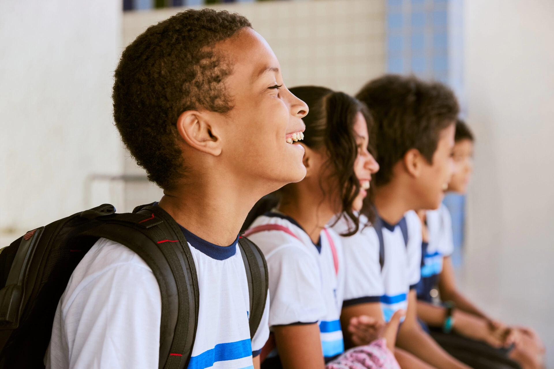 Quatro alunos usando uniformes e mochilas iguais sentam-se em uma fileira, sorrindo e olhando para a esquerda em um ambiente escolar ao ar livre - uma cena inspirada na dedicação vista nos programas educacionais do Plano Anual Instituto Ayrton Senna.