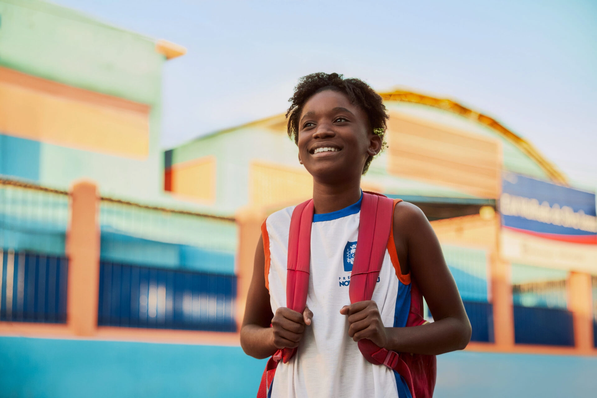 Um jovem sorridente, vestindo uma camiseta sem mangas branca e azul e uma mochila vermelha, está ao ar livre em frente a prédios coloridos, incorporando o espírito do Plano Anual Instituto Ayrton Senna.