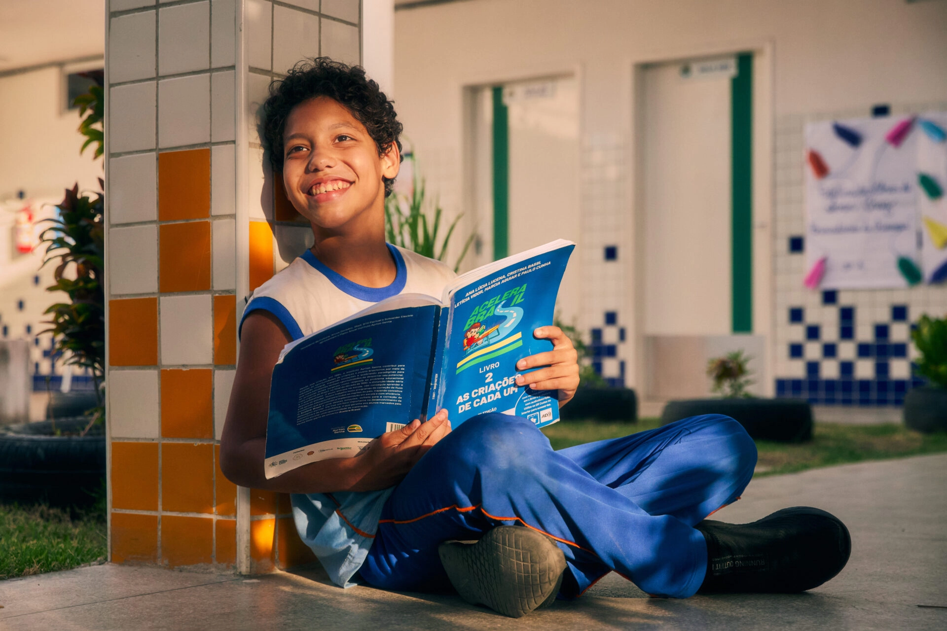 Um menino com uniforme escolar senta-se no chão ao ar livre, encostado em um pilar, sorrindo enquanto lê um livro didático do Plano Anual Instituto Ayrton Senna.