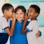 Três crianças estão juntas; dois meninos sussurram nos ouvidos de um menino sorridente no centro, com um quadro de avisos da sala de aula exibindo atividades do Plano Anual Instituto Ayrton Senna ao fundo.