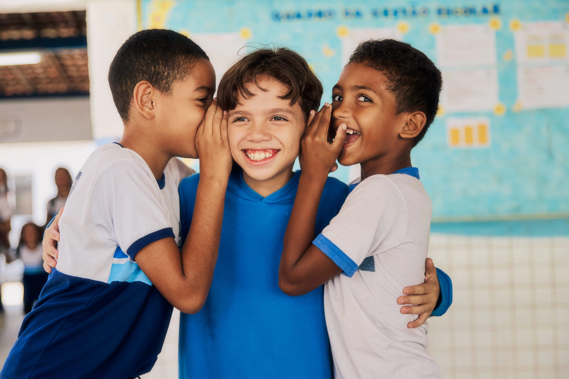 Três crianças estão juntas; dois meninos sussurram nos ouvidos de um menino sorridente no centro, com um quadro de avisos da sala de aula exibindo atividades do Plano Anual Instituto Ayrton Senna ao fundo.