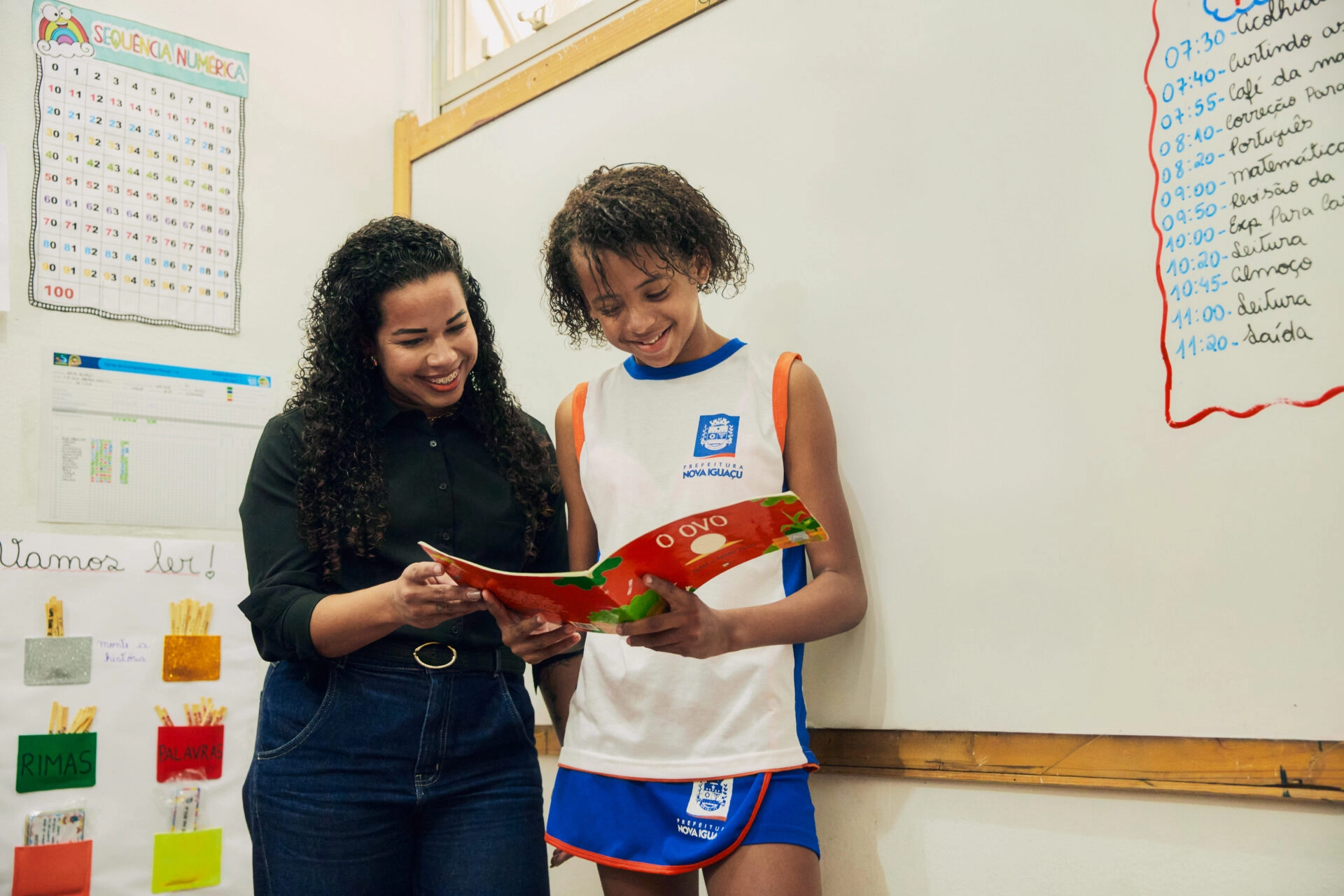 Um professor e um aluno estão ao lado de um quadro branco, sorrindo enquanto olham juntos para um livro vermelho em uma sala de aula, discutindo o Plano Anual Instituto Ayrton Senna.